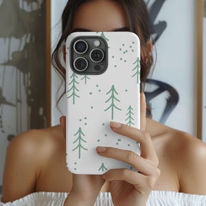 May include: A white smartphone case featuring a repeating pattern of sage green line-drawn pine trees and small dots. The case has a large rectangular cutout at the top left corner for the camera lenses and is held in a vertical orientation.