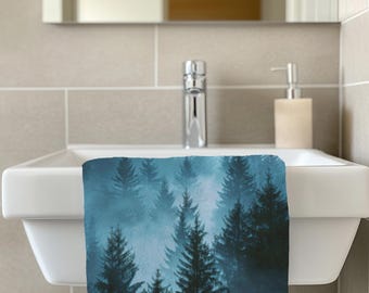 May include: A blue and white hand towel featuring a misty forest scene with tall evergreen trees. The towel is hanging over a white sink in a bathroom setting. A chrome faucet and a soap dispenser are also visible.