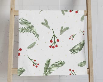 May include: A white towel featuring a repeating pattern of green pine needles and small red berries, accented with tiny gold dots scattered across the fabric. The design is printed on a rectangular textile surface with a clean, simple aesthetic.