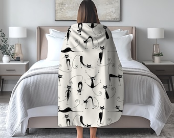May include: A cream-coloured blanket with a repeating pattern of black cat illustrations in various poses. The cats have long tails and are set against a backdrop of musical notes and swirling lines. The blanket is draped over a bed.
