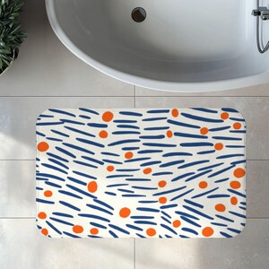 May include: A rectangular bath mat featuring an off-white background with a repeating pattern of short, horizontal dark blue brushstroke lines and scattered solid orange circles distributed across the entire surface of the mat.