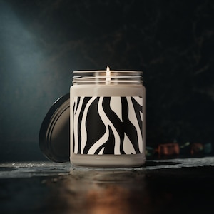 May include: A lit, gray candle in a clear glass jar with a black and white zebra print label. The lid is off to the side. The candle sits on a dark, reflective surface.