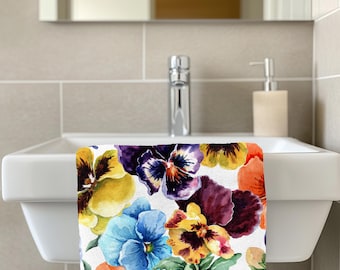 May include: A colorful hand towel featuring a watercolor floral pattern of pansies in shades of yellow, purple, blue, and orange. The towel is displayed on a white sink in a bathroom setting.
