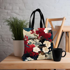 May include: A tote bag with black handles featuring a Japanese-inspired design. The artwork depicts red and white flowers on a dark blue background with stylized waves and a tree branch. The bag is on a wooden table.