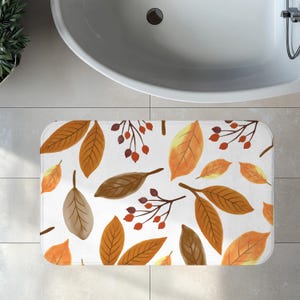 Op de afbeelding: Rechthoekige witte badmat met een aquarelpatroon van herfstbladeren in oranje, bruin en geel, en rode bessen. De mat ligt voor een witte badkuip.