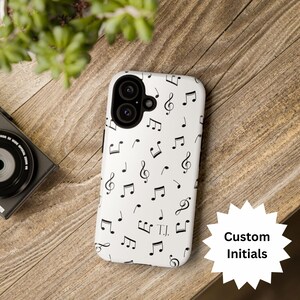 Puede incluir: Funda de teléfono blanca con un patrón de notas musicales negras. La funda tiene un recorte para la cámara y el texto "Custom Initials" en una estrella blanca. La funda del teléfono está sobre una superficie de madera.