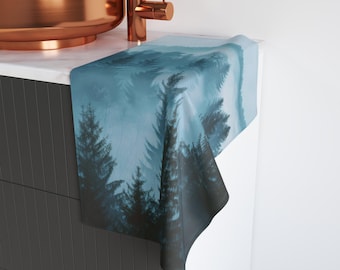 May include: A teal and blue forest scene printed on a rectangular towel. The design features silhouetted evergreen trees in a misty landscape. The towel is draped over a white countertop next to a copper sink.