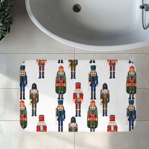 May include: A rectangular bath mat featuring a repeating pattern of nutcracker figures on a white background. The nutcrackers are depicted in various uniforms of red, blue, and green, wearing tall hats and holding small gold-colored instruments.