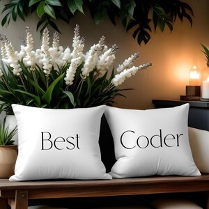 Könnte beinhalten: Zwei weiße Kissen mit den Wörtern "Best Coder" in Schwarz aufgedruckt. Die Kissen liegen auf einer Holzbank, im Hintergrund ein Blumenarrangement und Kerzen.