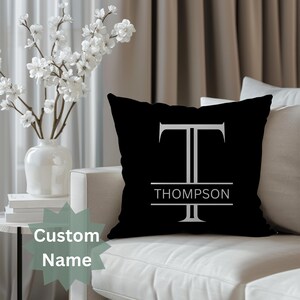 May include: Black cushion featuring a white letter "T" and the name "THOMPSON". The cushion is placed on a white sofa. The words "Custom Name" are in a green starburst.
