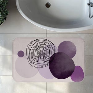 May include: Rectangular bath mat with a light purple background and abstract design. The design includes a black spiral and several purple circles of varying sizes and shades. The mat is placed in front of a white bathtub.