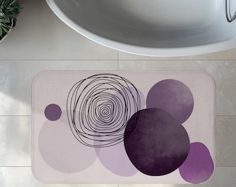 May include: Rectangular bath mat with a light purple background and abstract design. The design includes a black spiral and several purple circles of varying sizes and shades. The mat is placed in front of a white bathtub.