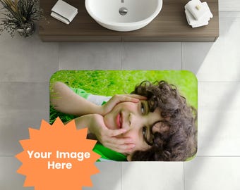 May include: A rectangular bath mat featuring a vibrant image of a child lying on green grass. The child is smiling and resting their face on their hands. An orange starburst graphic with the text "Your Image Here" is in the foreground.