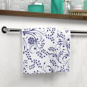 Pode incluir: Toalha branca com um padrão floral azul pendurada numa barra de toalha cromada. A toalha apresenta um design repetido de flores e vinhas estilizadas. O acessório de casa de banho é exibido contra uma parede de azulejos brancos.