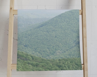 May include: A rectangular towel featuring a scenic mountain landscape covered in lush green trees. The towel is displayed on a light-colored wooden ladder-style rack against a white tiled wall.
