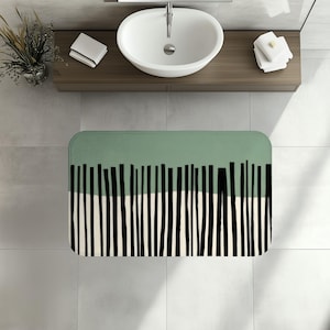 May include: A rectangular bath mat featuring a horizontal color-blocked design. The top section is sage green, while the bottom section is off-white. A series of vertical, uneven black lines of varying widths span across the center of the mat.