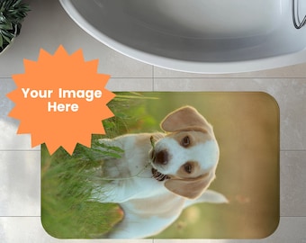 May include: A rectangular bath mat featuring a light brown and white puppy in a grassy field. The puppy is looking at the viewer. The mat has a rounded edge and is placed in front of a white bathtub.