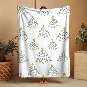 Puede incluir: Una manta blanca con un patrón repetitivo de árboles de Navidad dibujados con líneas de color verde oscuro, cada uno coronado con una estrella amarilla sólida. La manta se sostiene para mostrar la orientación vertical del diseño.