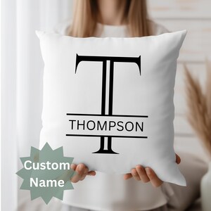 May include: White square cushion featuring a large black letter "T" and the name "THOMPSON" printed beneath. A custom name tag is in the lower left corner. The cushion is held up by a person.