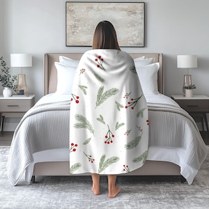 May include: A white throw blanket featuring a repeating pattern of green pine needles and clusters of red berries. The blanket is draped over the back of a person standing in front of a bed with white pillows and a gray quilted bedspread.