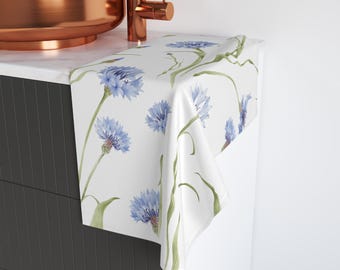 May include: White hand towel featuring a watercolor design of blue cornflowers and green stems. The floral pattern is repeated across the fabric. The towel is draped over a countertop, showcasing the design.