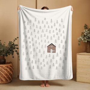 May include: A white fleece blanket featuring a repeating pattern of small, black, hand-drawn pine trees. Centered on the blanket is a single, detailed illustration of a brown cabin with a white door and windows, surrounded by the forest pattern.