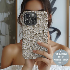 May include: A phone case featuring a floral pattern of small white flowers with yellow centers against a light beige background. The case has a black camera module and the text "PRINTED DESIGN NOT 3D NOT REAL STAINED GLASS".