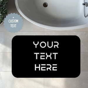 May include: A black rectangular bath mat with rounded corners. The mat features the words "YOUR TEXT HERE" in white, block letters. A light blue, scalloped-edged sticker with the words "CUSTOM TEXT" is in the upper left corner. A white bathtub is in the background.
