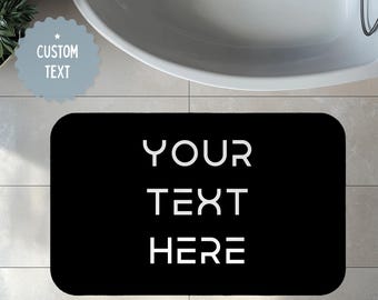 May include: A black rectangular bath mat with rounded corners. The mat features the words "YOUR TEXT HERE" in white, block letters. A light blue, scalloped-edged sticker with the words "CUSTOM TEXT" is in the upper left corner. A white bathtub is in the background.