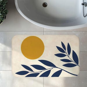 May include: A rectangular bath mat featuring a cream-coloured background with a solid mustard yellow circle in the upper left corner and a dark navy blue botanical branch with multiple pointed leaves extending across the lower portion of the mat.