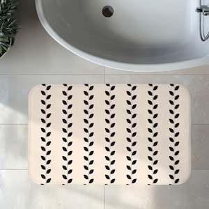 May include: A rectangular bath mat featuring a cream-colored base with a repeating pattern of vertical black vine-like lines. Each line consists of small, uniform black leaves arranged in a symmetrical, alternating sequence across the surface.