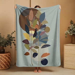 以下が含まれることがあります： 抽象的な植物のデザインが施された、柔らかい水色のブランケット。茶色、緑、オレンジ、青の色合いの様式化された葉が、茶色と青の円の背景に配置されています。ブランケットは人が持っています。