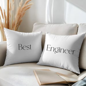 May include: Two white cushions on a beige sofa. One cushion says "Best" in black script, the other says "Engineer". An open book rests on the sofa. Dried decorative plants are in the background.