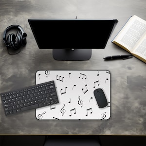 May include: A computer desk setup featuring a black monitor, keyboard, mouse, and headphones. A white mousepad with black musical notes is in the center. An open book and pen are also on the desk. The desk has a gray, textured surface.