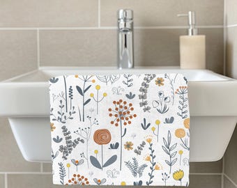 May include: White hand towel with a floral pattern featuring orange, yellow, and gray botanical illustrations. The towel is hanging on a white sink in a bathroom setting. The design includes various flowers and leaves.
