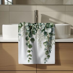 May include: A white hand towel featuring a printed botanical design of cascading eucalyptus branches and small white flowers. The greenery displays various shades of sage and forest green, creating a natural pattern across the fabric surface.
