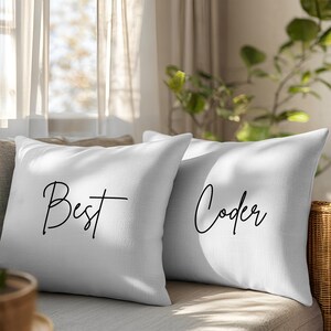 Könnte beinhalten: Zwei weiße Kissen mit schwarzer Schrift. Auf einem Kissen steht "Best" und auf dem anderen "Coder". Die Kissen liegen auf einem hellen Sofa, mit einem Fenster und Pflanzen im Hintergrund.