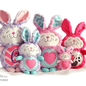 May include: Four plush bunny toys in different sizes, with pink, purple and blue fur. The largest bunny has a pink and white chevron pattern on its tummy and a pink letter C. The other bunnies have hearts on their tummies.