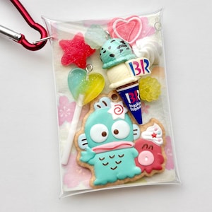 May include: A clear plastic pouch with a red carabiner clip, filled with colorful charms. Charms include a blue cartoon character cookie, ice cream cone, star, heart, and lollipop. The ice cream cone has the letters "BR" on it.