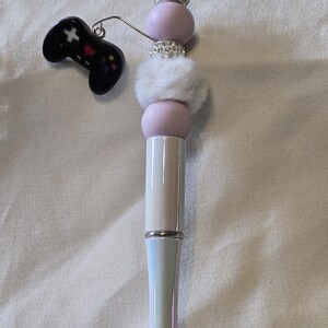 May include: A decorative pen with a black video game controller charm. The pen features a white and iridescent barrel, adorned with pink beads, faux fur, and sparkling rhinestones. The pen is designed for writing.