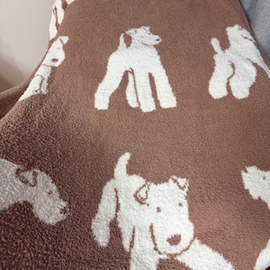 Op de afbeelding: Een bruine deken met een herhaald patroon van witte hondenillustraties. De honden worden in verschillende poses afgebeeld, met hun lichamen en kenmerken duidelijk omlijnd tegen de bruine achtergrond. De deken heeft een zacht, getextureerd uiterlijk.