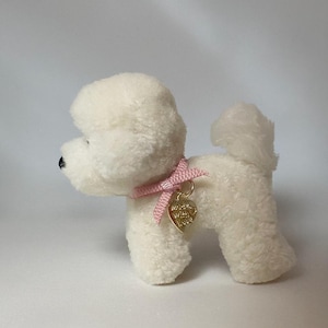 May include: A small, white plush dog with a pink ribbon collar and a gold heart-shaped tag. The tag has engraved text. The dog has a fluffy tail and a black nose. The plush toy is designed to resemble a Bichon Frise.