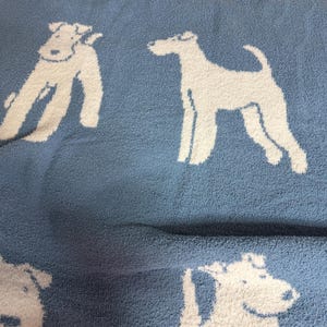 Puede incluir: Una manta azul claro con un patrón repetido de siluetas de perros blancos. Los perros están representados en diferentes poses, creando un diseño lúdico. La manta tiene un aspecto suave y texturizado.