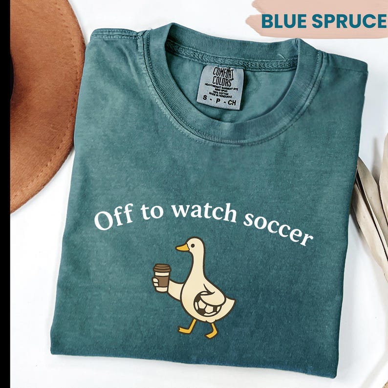 May include: Blue Spruce Comfort Colors t-shirt with the text "Off to watch soccer" and a cartoon duck holding a coffee cup. The duck has a soccer ball design on its body. The shirt is folded.