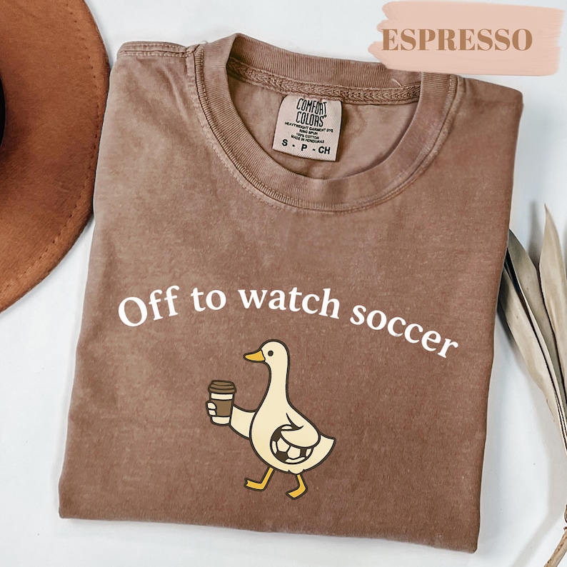 May include: Espresso-colored t-shirt with the text "Off to watch soccer." A cartoon duck holding a coffee cup and a soccer ball is printed on the front. The shirt is a Comfort Colors brand, size small.