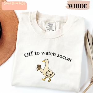 May include: Cream-colored t-shirt with the phrase "Off to watch soccer" and a cartoon duck holding a coffee cup. The shirt is made of a soft material and has a tag that says "Comfort Colors".