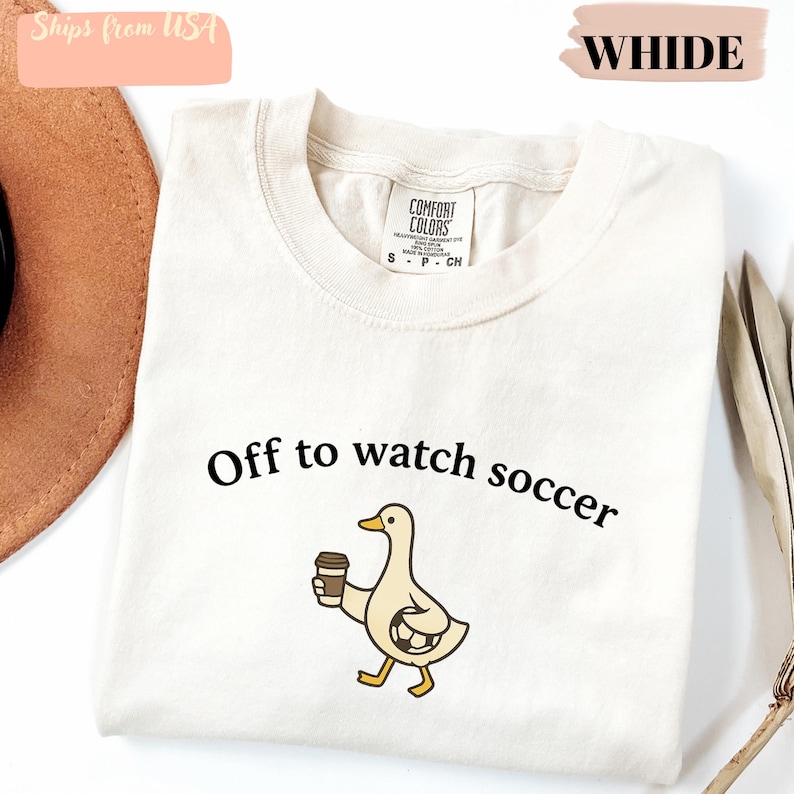 May include: Cream-colored t-shirt with the phrase "Off to watch soccer" and a cartoon duck holding a coffee cup. The shirt is made of a soft material and has a tag that says "Comfort Colors".