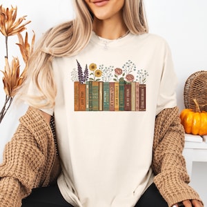 May include: Cream-colored t-shirt featuring a graphic of a row of books with flowers on top. The books have titles like "Love Story" and "Poetry". The flowers include sunflowers, roses, and lavender. The shirt has a relaxed fit.