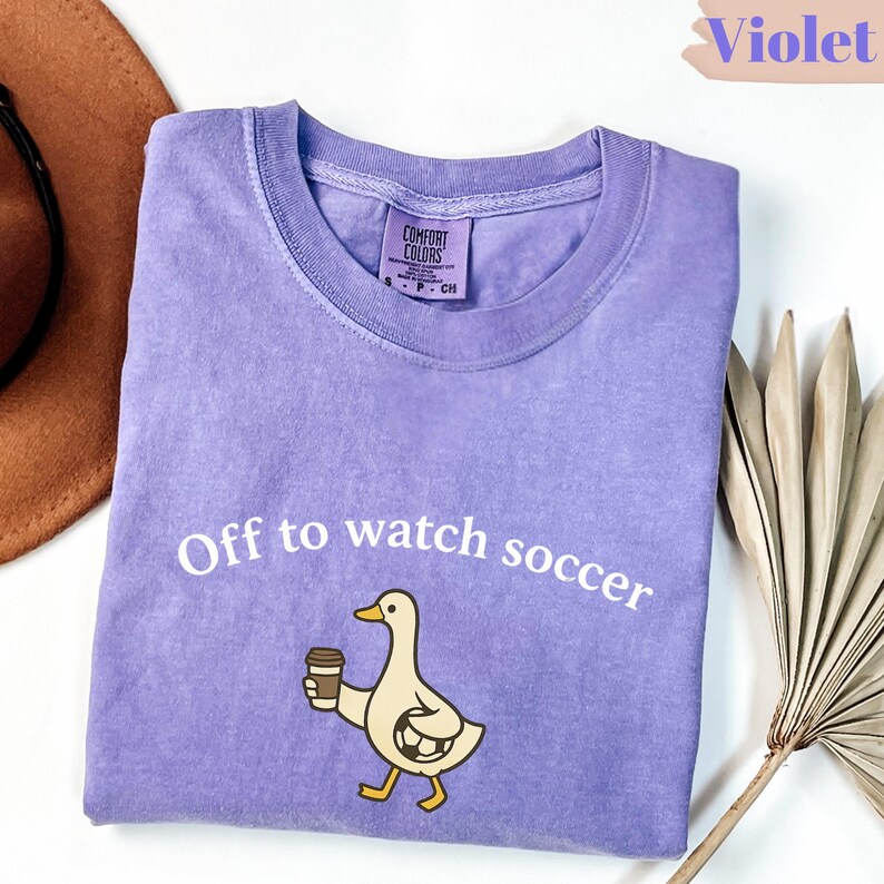 May include: Violet Comfort Colors t-shirt with the text "Off to watch soccer" and a cartoon duck holding a coffee cup. The duck is wearing a soccer ball on its body. The shirt is a light purple color.