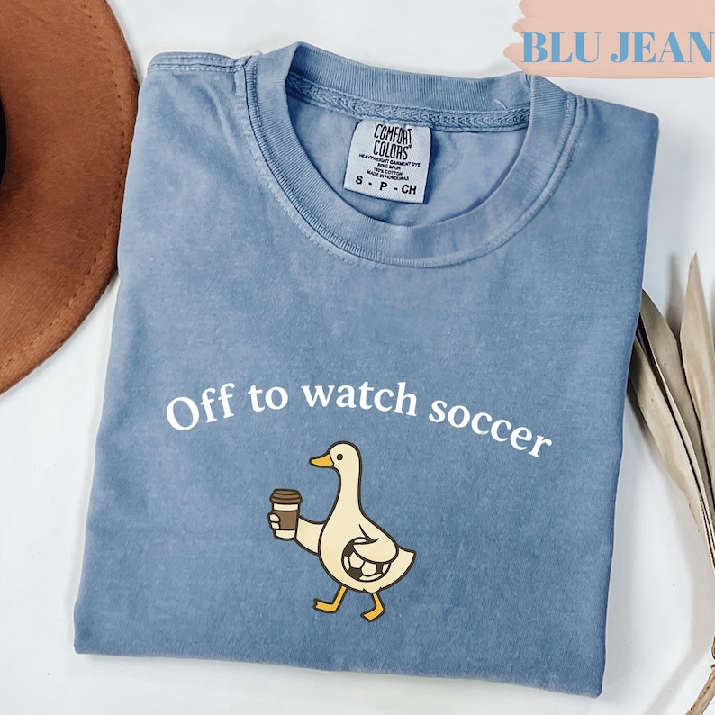 May include: A blue Comfort Colors t-shirt with the text "Off to watch soccer." The shirt features a cartoon duck holding a coffee cup and a soccer ball. The shirt is a light blue color.